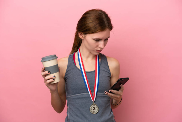 Joven mujer inglesa con medallas aisladas sobre fondo rosa sosteniendo café para llevar y un móvil - Foto, Imagen