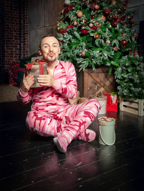 Cheerful funny happy man in pink sleepwear sit near decorated fir tree with Christmas present and sending kiss - Photo, Image