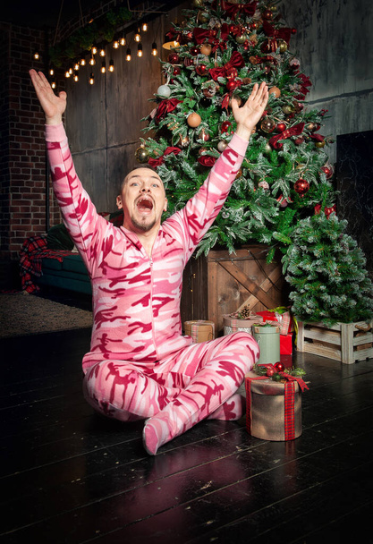 Cheerful surprised funny man in pink sleepwear with hands up sitting on the floor near decorated fir tree and Christmas present - Photo, Image