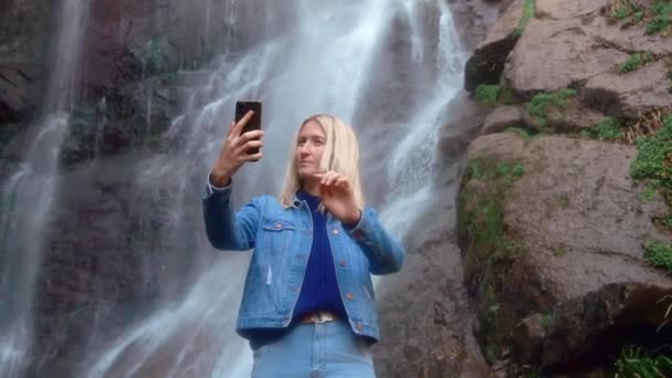 woman holds smartphone and takes pictures of herself standing near waterfall - Filmagem, Vídeo