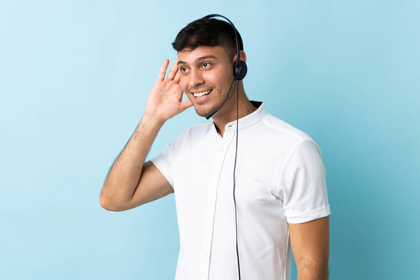 Telemarketer Colombiaanse man werken met een headset over geïsoleerde achtergrond luisteren naar iets door de hand op het oor - Foto, afbeelding