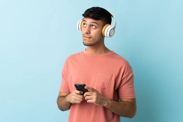 Молодой колумбиец изолирован на синем фоне, слушает музыку на мобильном телефоне и думает: - Фото, изображение