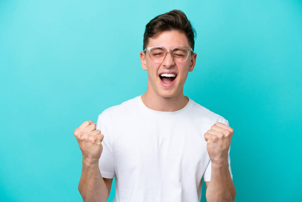 Genç ve yakışıklı Brezilyalı bir adam mavi arka planda gözlüklü ve zaferi kutluyor. - Fotoğraf, Görsel