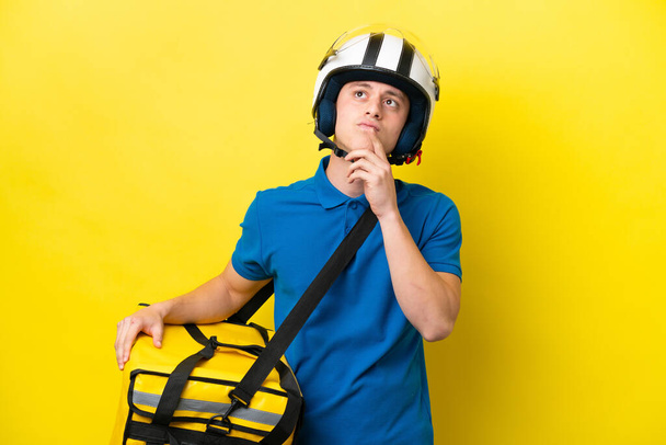 Young Brazilian man with thermal backpack isolated on yellow background having doubts while looking up - 写真・画像