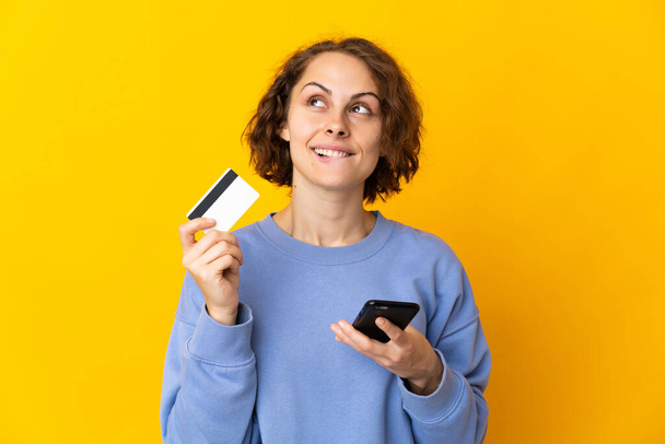 Молода англійська жінка ізольована на рожевому фоні, купуючи мобільний телефон з кредитною карткою під час мислення. - Фото, зображення