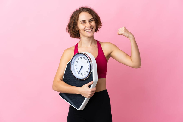 Young English woman isolated on pink background holding a weighing machine and doing strong gesture - Foto, immagini