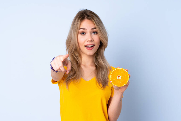 Teenager Russian girl holding an orange isolated on blue background points finger at you with a confident expression - Фото, изображение