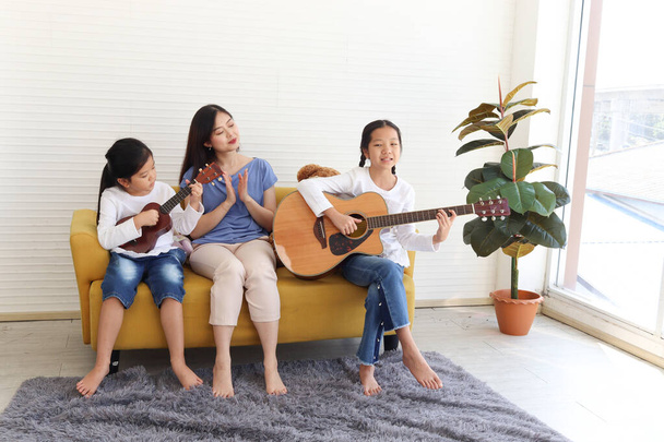 Дві щасливі доньки і мати співають пісню і грають на гітарі укулеле, сидячи на дивані у вітальні, мама і діти проводять разом особливий момент.. - Фото, зображення