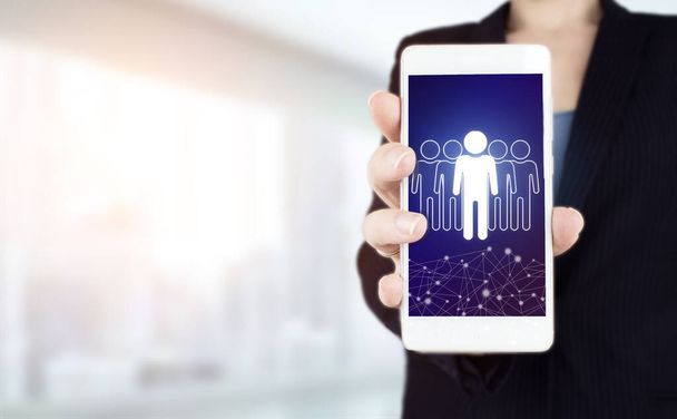 Human resources and team management concept. Hand hold white smartphone with digital hologram Human, Leader sign on light blurred background. Community, unity, people and support concept - Photo, Image