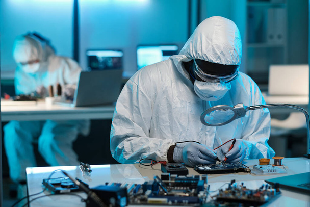 Technician in protective coveralls, eyeglasses, gloves and respirator repairing circuit board - Foto, imagen