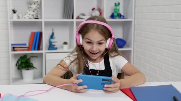 onnellinen yllättynyt lapsi kuulokkeissa katsomassa videota puhelimessa luokkahuoneessa, estudy - Materiaali, video