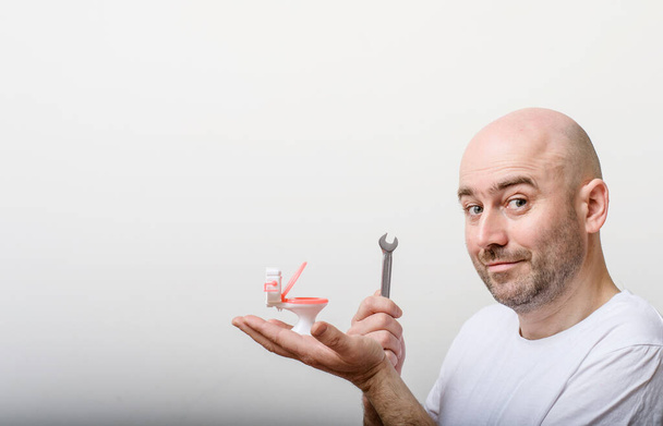 Portrait of confused bald man with toy flush toilet bowl on white background. Repair by plumber. Plumbing problems. - Фото, изображение