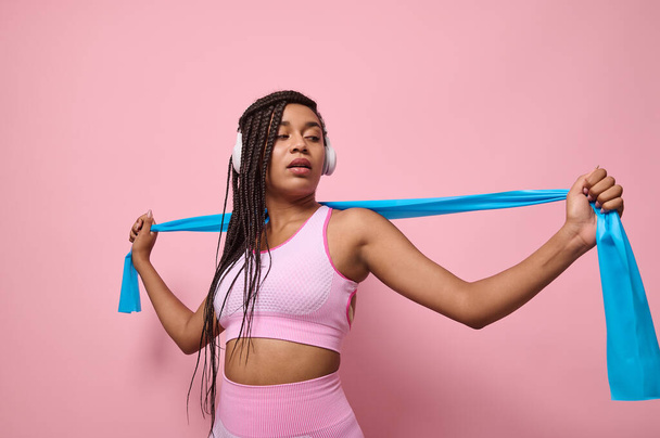 African sportswoman with wireless headphones performs exercises with resistance band. People, sport and fitness. Young woman training with elastic strap on pink background. Active lifestyle concept - Fotó, kép