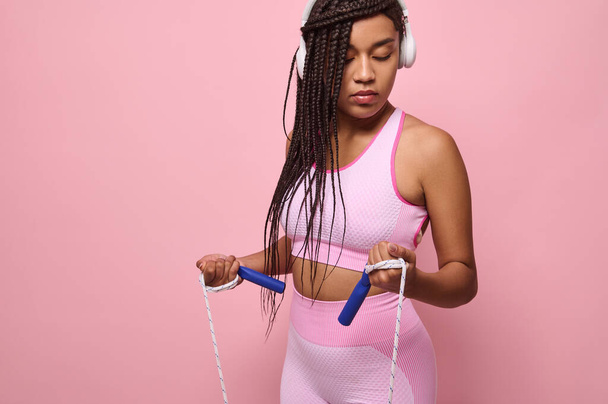 Horizontal portrait of a sporty African determined woman in pink sportswear wearing wireless headphones, exercising with skipping rope, isolated over colored background with copy space - Photo, Image