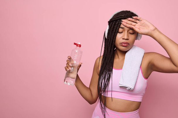 Moe uitgeput dorstige fitnessvrouw met draadloze hoofdtelefoon en een handdoek op haar schouders die een fles water vasthoudt, ontspant na zware training, poseert tegen een roze achtergrond met kopieerruimte - Foto, afbeelding