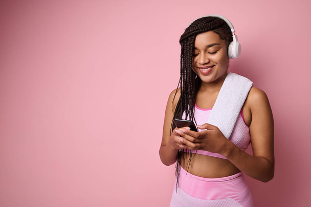 Attraktive afrikanische Sportlerin in rosa Trainingsanzug und drahtlosen Kopfhörern, surft mit Smartphone im Internet, wischt, surft, isoliert über rosafarbenem Hintergrund - Foto, Bild