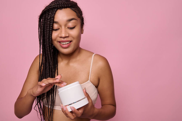 Smiling African woman in underclothes posing with a jar with beauty cosmetic product and applying it, posing against a pink colored background - Photo, Image