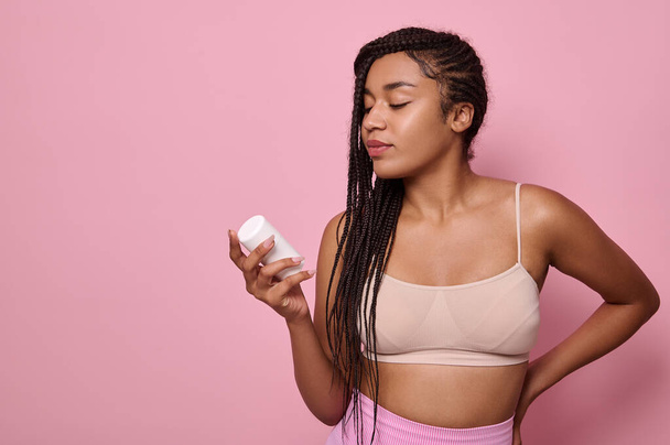 Beautiful African woman with stylish afro dreadlocks in underclothes holding a bottle with beauty cosmetic product, isolated over pink background with copy space for advertisement - Photo, image