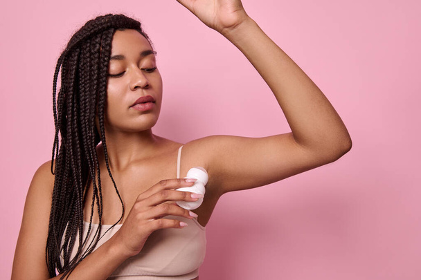 Close-up isolated beauty portrait on pink background of African young woman with dreadlocks in beige underwear applying antiperspirant under the armpits. Purity, hygiene, body care concept. Copy space - Zdjęcie, obraz