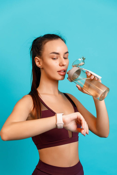 Spor giyimli güzel esmer kadın elinde bir şişe suyla dikilip mavi arka plandaki saatine bakıyor. Metin için boşluk - Fotoğraf, Görsel