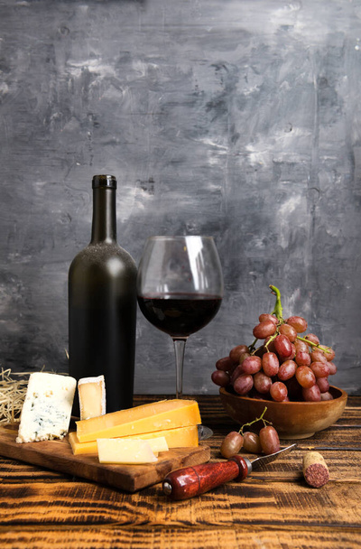 Red wine in a dusty bottle and glass. Grapes with cheese on a wooden table. Space for text. Gray background. - Foto, immagini