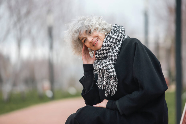 A beautiful mature grey haired lonely woman is sitting on a bench in a cold autumn park - Photo, Image