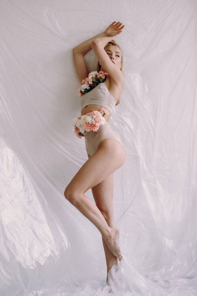Beautiful blonde in beige underwear with carnations. Body care, beautiful figure of a girl, flowers growing from a person. - Fotoğraf, Görsel