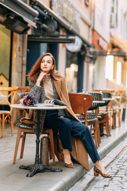 イスタンブールの古い通りを女が歩いている。ガラタ塔を背景に茶色のコートを着た少女の肖像画. - 写真・画像