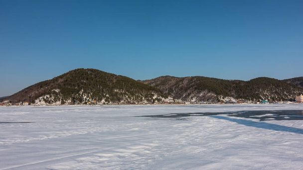 Het bevroren meer is bedekt met sneeuw. Gebieden met helder ijs zijn zichtbaar. Kustbeboste bergketen tegen de blauwe lucht. Baikal. - Foto, afbeelding