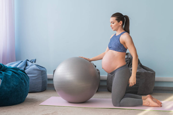 Pregnant european woman performs exercises with fitness ball. Expectant mother is doing yoga in the 3rd trimester. Girl in the classroom in preparation for childbirth. - Fotó, kép