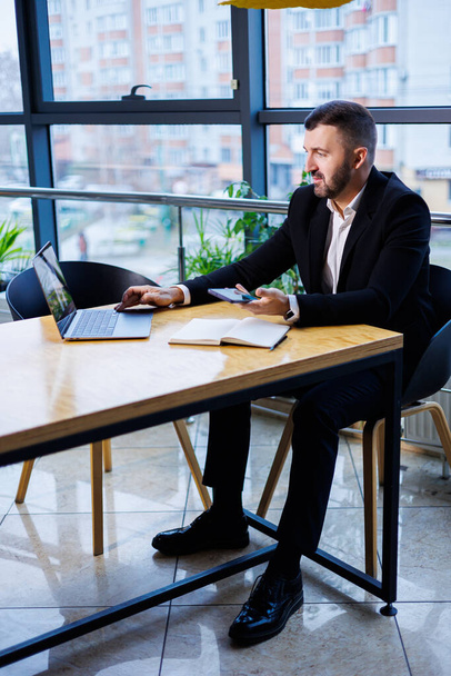 Portrait of a stylish young business man, in a large modern office high on an upper floor, looking into a laptop. Business man at freelancer finance office desk - Foto, Imagen