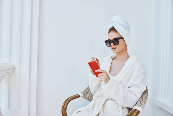 vrouw met een telefoon in een witte badjas zit op een balkon in een hotel - Foto, afbeelding