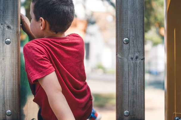 7-jarige jongen met rood t-shirt zittend op de glijbaan in de speeltuin met zijn rug naar de camera en ontspannend - Foto, afbeelding
