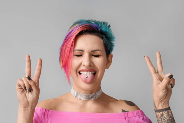 Cool woman with unusual hair showing victory gesture on grey background - Zdjęcie, obraz