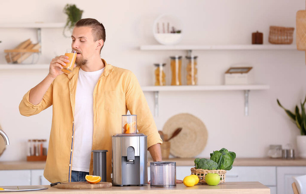 Young man with glass of fresh fruit juice near modern juicer in kitchen - Φωτογραφία, εικόνα