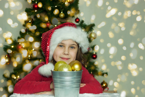 Portrait of a girl with Christmas tree toys. Smiling girl in Santa hat sits next to Christmas tree toys - Foto, afbeelding
