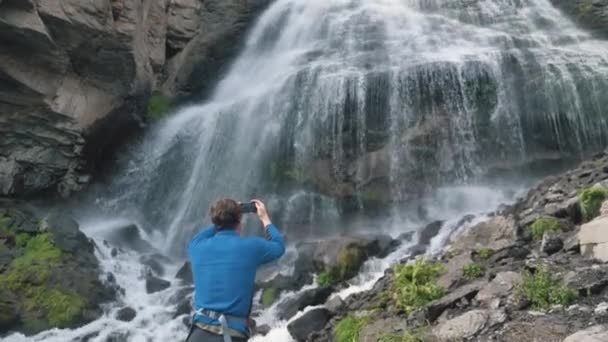 El hombre sostiene el teléfono y hace video de pie cerca de la cascada - Metraje, vídeo
