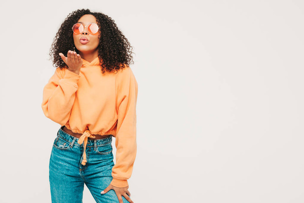 Beautiful black woman with afro curls hairstyle.Smiling model in orange hoodie and trendy jeans clothes. Sexy carefree female posing on white background in studio. Gives air kiss - Fotografie, Obrázek