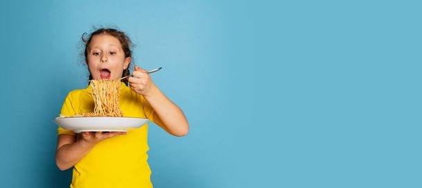 Cute little girl, emotive kid eating delicious Italian pasta isolated on blue studio background. World pasta day - 写真・画像