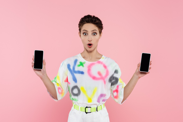 surprised brunette woman showing smartphones with blank screen isolated on pink - Фото, изображение