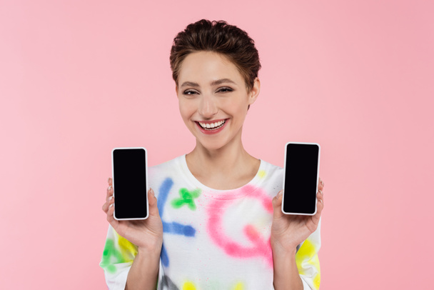 young brunette woman showing mobile phones while smiling at camera isolated on pink - Foto, Imagem