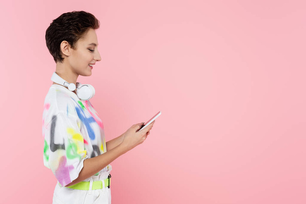 side view of smiling woman with wireless headphones on neck using cellphone isolated on pink - Foto, afbeelding