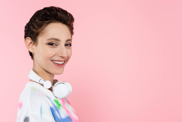 cheerful woman with wireless headphones on neck looking at camera isolated on pink - Foto, Bild