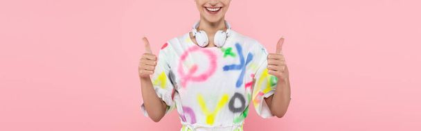 oříznutý pohled na šťastnou ženu se sluchátky na krku ukazující palce izolované na růžové, banner - Fotografie, Obrázek