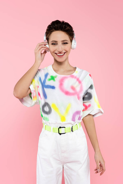 happy woman in stylish clothes and headphones looking at camera isolated on pink - Photo, Image