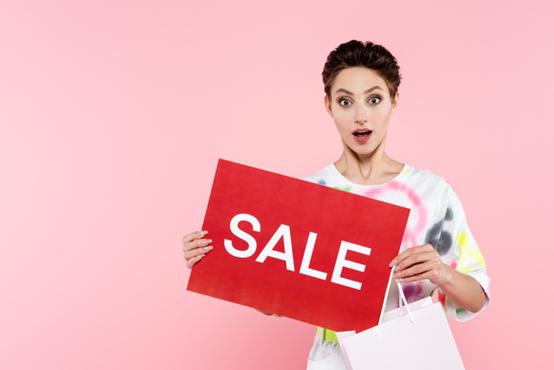 thrilled woman with shopping bag and red card with sale lettering looking at camera isolated on pink - Photo, image