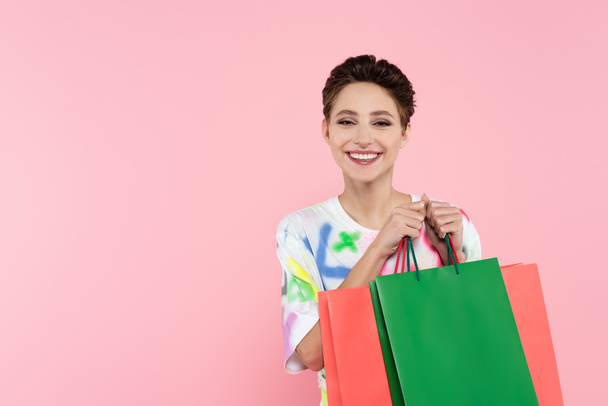 potěšená brunetka žena usmívá na kameru, zatímco drží nákupní tašky izolované na růžové - Fotografie, Obrázek
