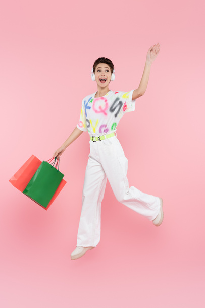 vista completa de la mujer alegre en auriculares levitando con bolsas de compras mientras agita la mano en rosa - Foto, Imagen