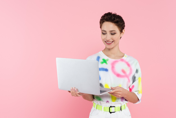 young smiling woman in stylish clothes standing with laptop isolated on pink - Foto, Bild