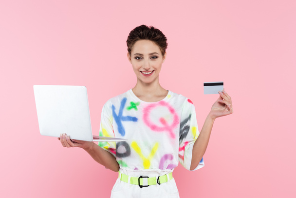 glückliche brünette Frau mit Laptop und Kreditkarte schaut in die Kamera isoliert auf rosa - Foto, Bild